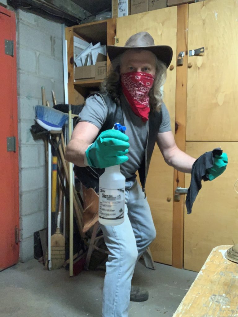 Wearing a cowboy hat and a red bandana over his mouth, a long-haired man poses with cleaning supplies drawn like a gun