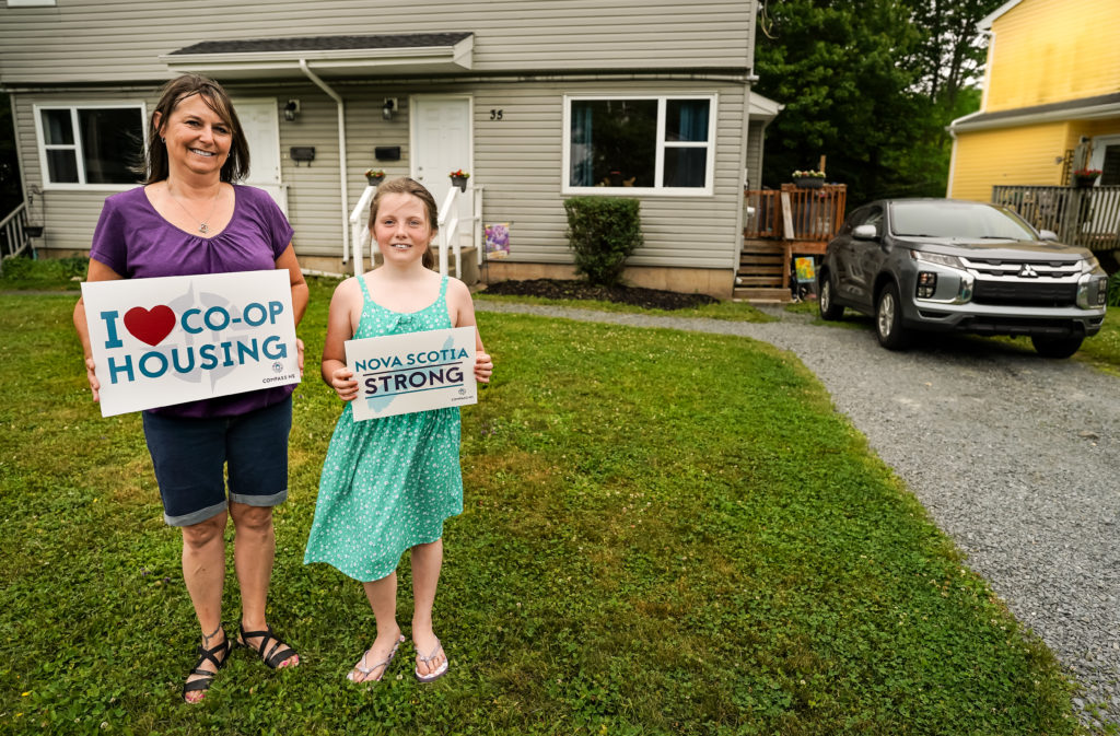 A mother and daughter stand smiling on the lawn of their co-op home. They hold signs saying "We love co-op housing" and "Halifax co-ops"