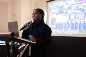 A woman speaks with a slideshow behind her.