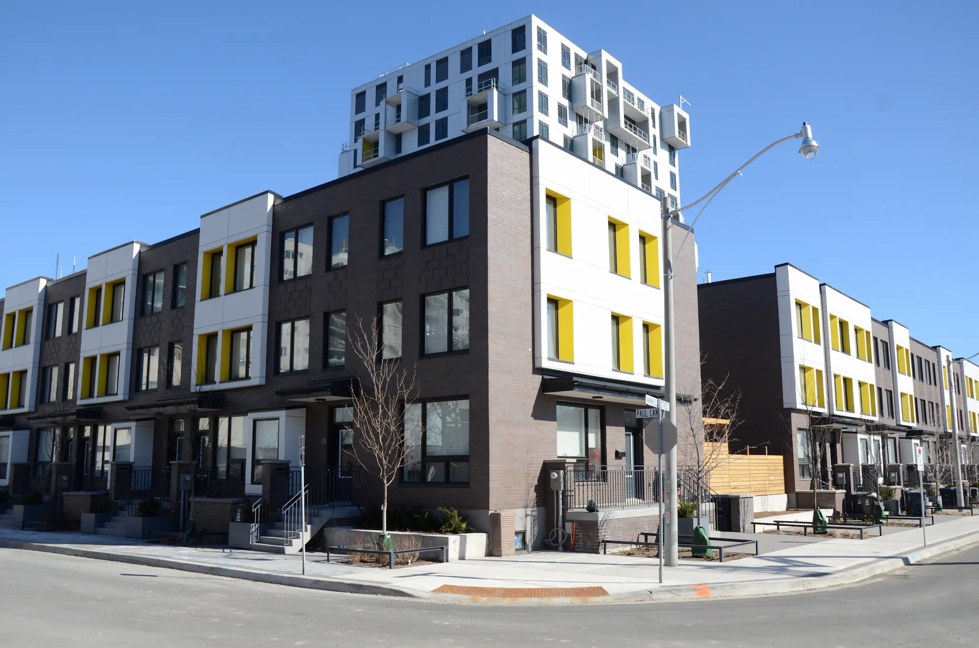 Modern townhouse buildings with yellow window accents and a multi-story structure on top, exemplifying co-operative housing, are situated on a clear, sunny day at a street corner.