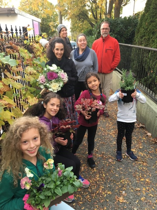 Un groupe d'adultes et d'enfants se tiennent à l'extérieur sur un trottoir, tenant des plantes en pot et des fleurs devant des feuilles d'automne, célébrant l'habitation coopérative avec la FHCC.