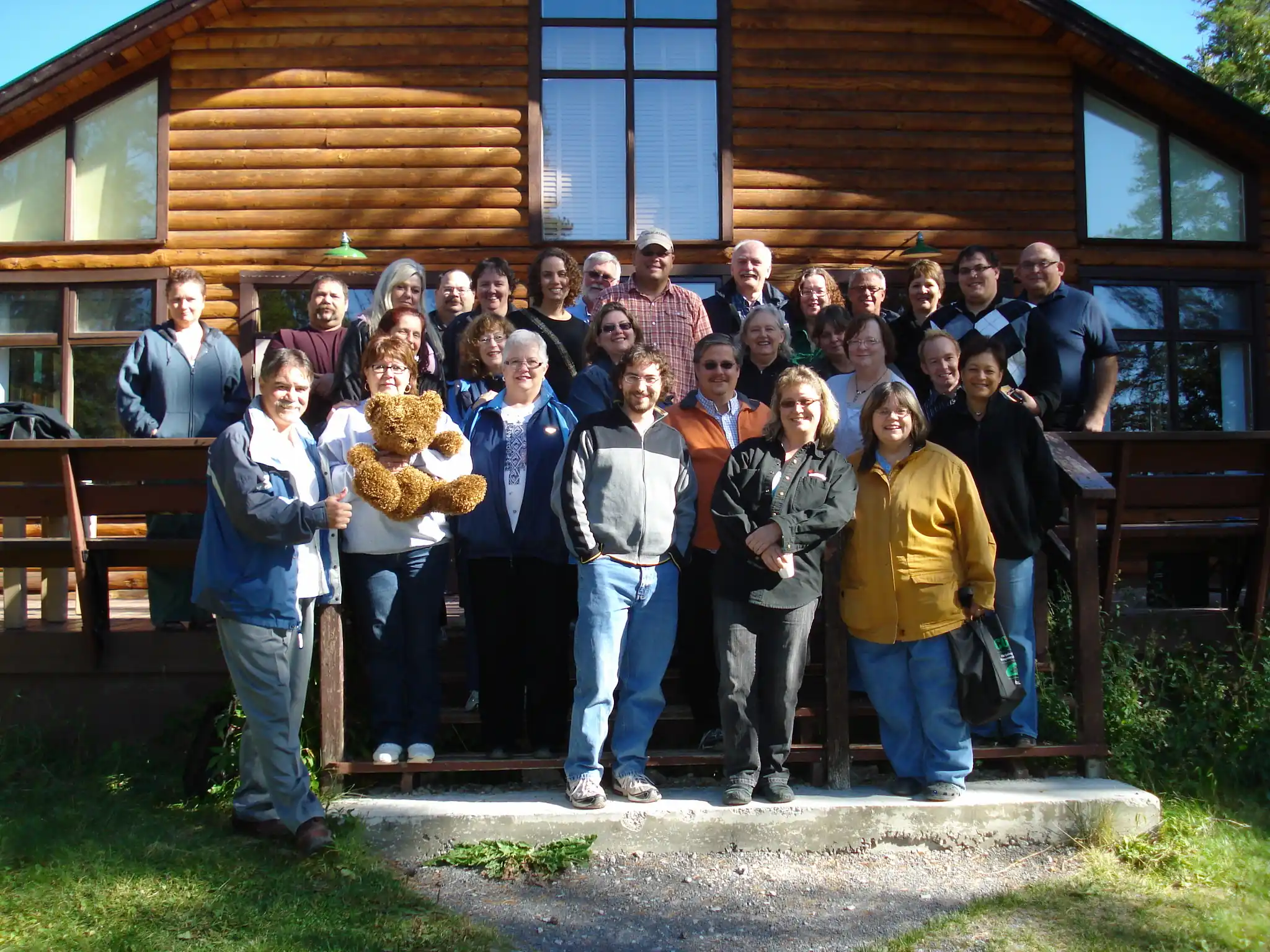 Un groupe d'adultes pose pour une photo sur les marches d'une cabane en rondins, avec une personne tenant un ours en peluche, célébrant l'habitat coopératif par une journée ensoleillée.
