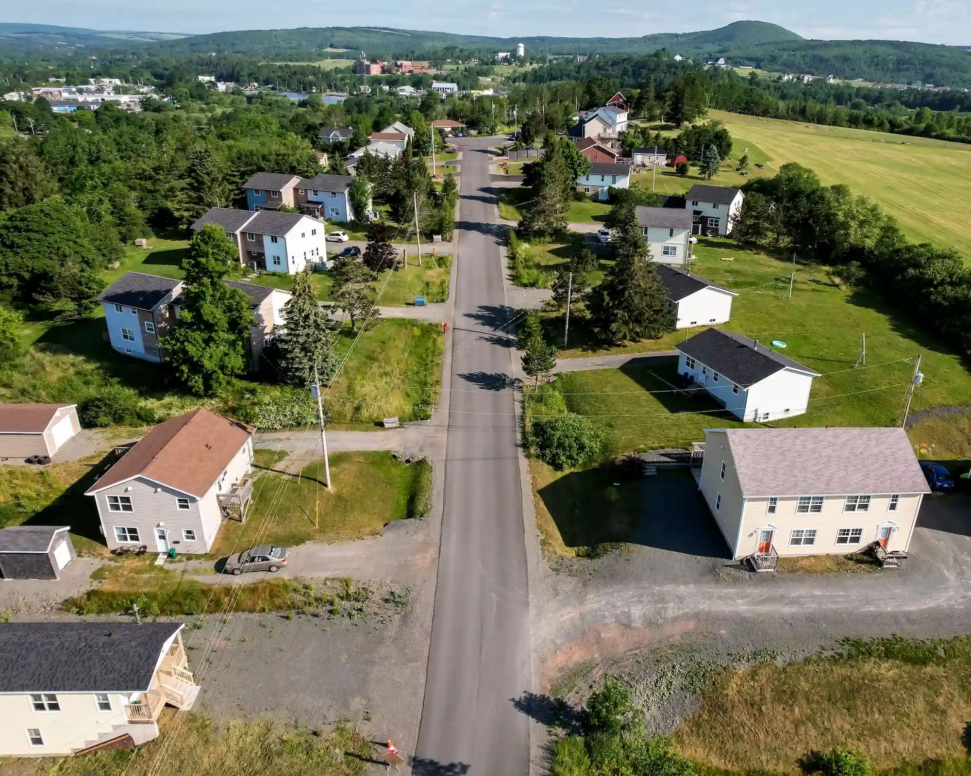 Vue aérienne d'une rue de banlieue tranquille bordée de coopératives d'habitation, de pelouses vertes et d'arbres, avec des champs et des collines visibles à l'arrière-plan - reflétant la vision de la FHCC pour des communautés dynamiques et durables.