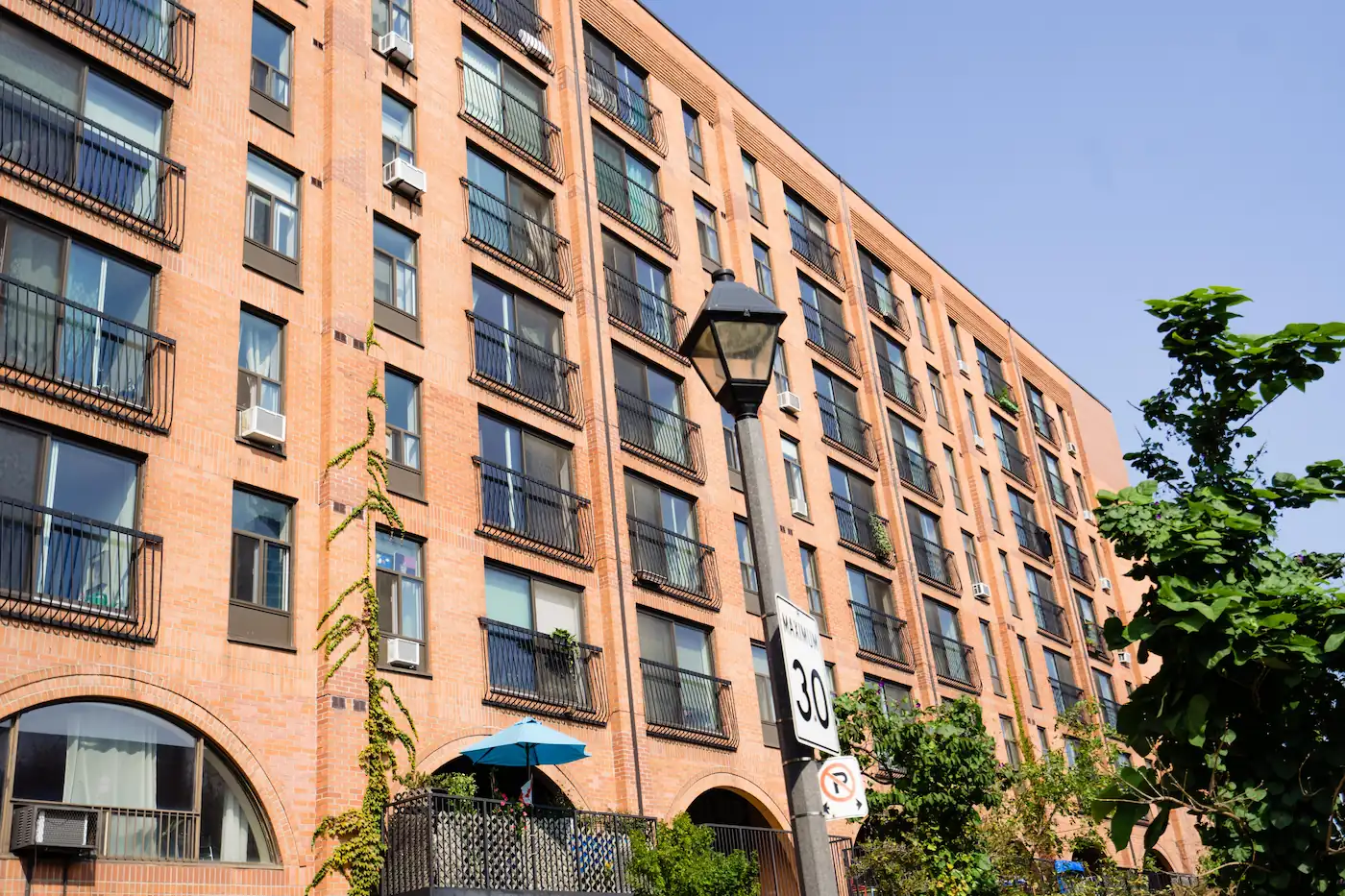 A multi-story brick co-operative housing apartment building with balconies and large windows, a 30 km/h speed limit sign, and greenery in the foreground under a clear sky.