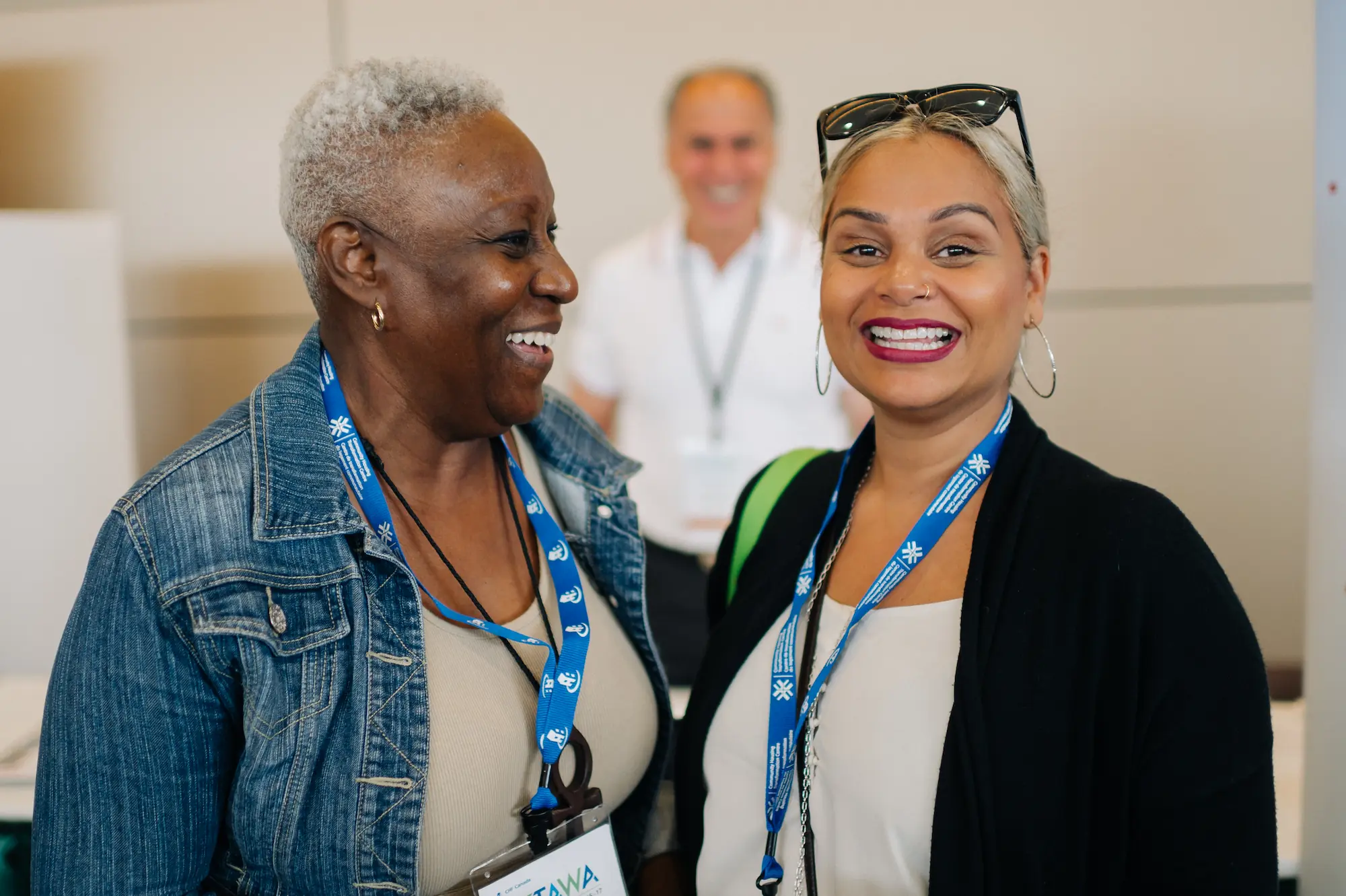 Deux femmes portant des cordons de conférence sourient à l'appareil photo, représentant l'habitation coopérative lors d'un événement de la FHCC, avec un homme souriant dans l'arrière-plan flou.