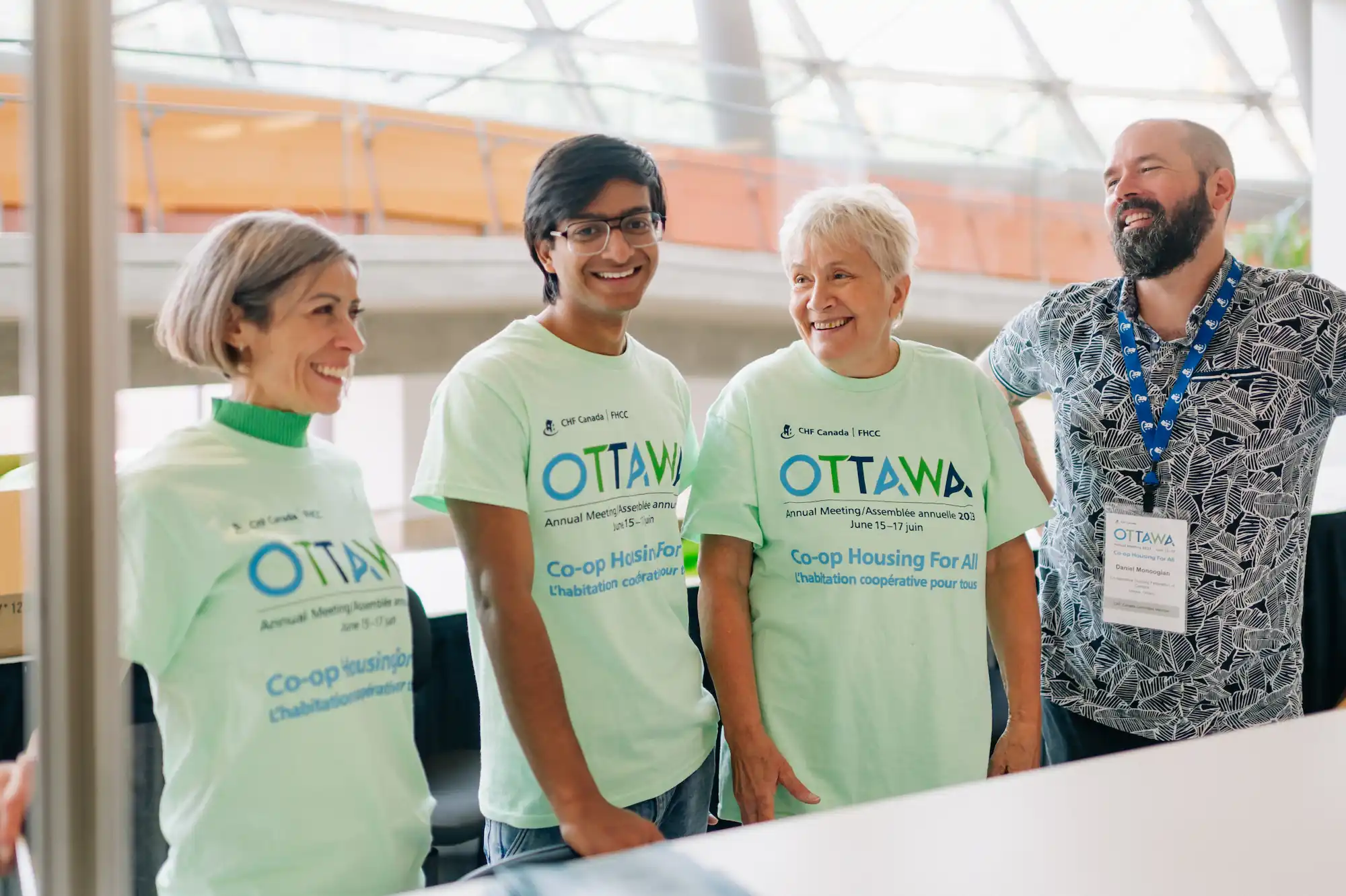 Quatre personnes se tiennent ensemble à l'intérieur, souriantes et portant des t-shirts vert pâle "Ottawa Co-op Housing For All" lors d'un événement sur l'habitation coopérative. Une personne porte un insigne nominatif et un cordon, montrant qu'elle appuie les initiatives de logement de la FHCC.