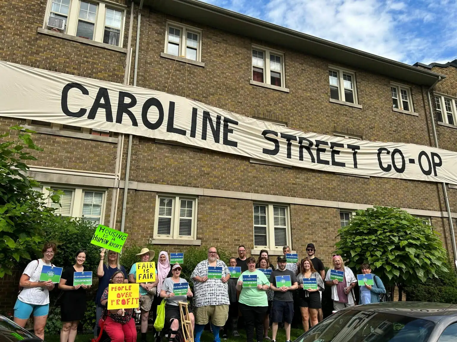 Un groupe de personnes se tient devant un bâtiment en briques avec une grande bannière indiquant "CAROLINE STREET CO-OP", tenant des pancartes qui font la promotion de l'habitation coopérative et de la défense des droits, montrant leur appui à des organismes comme la FHCC.