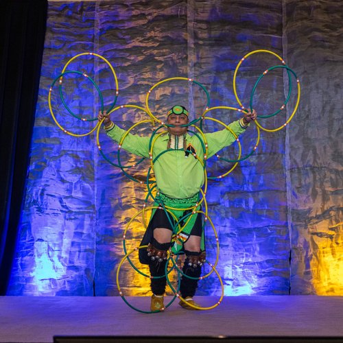 A performer in bright clothing stands on stage holding multiple colorful hoops, their vibrant energy reminiscent of the unity found in Co-operative Housing, against a textured backdrop lit with blue and yellow lights.