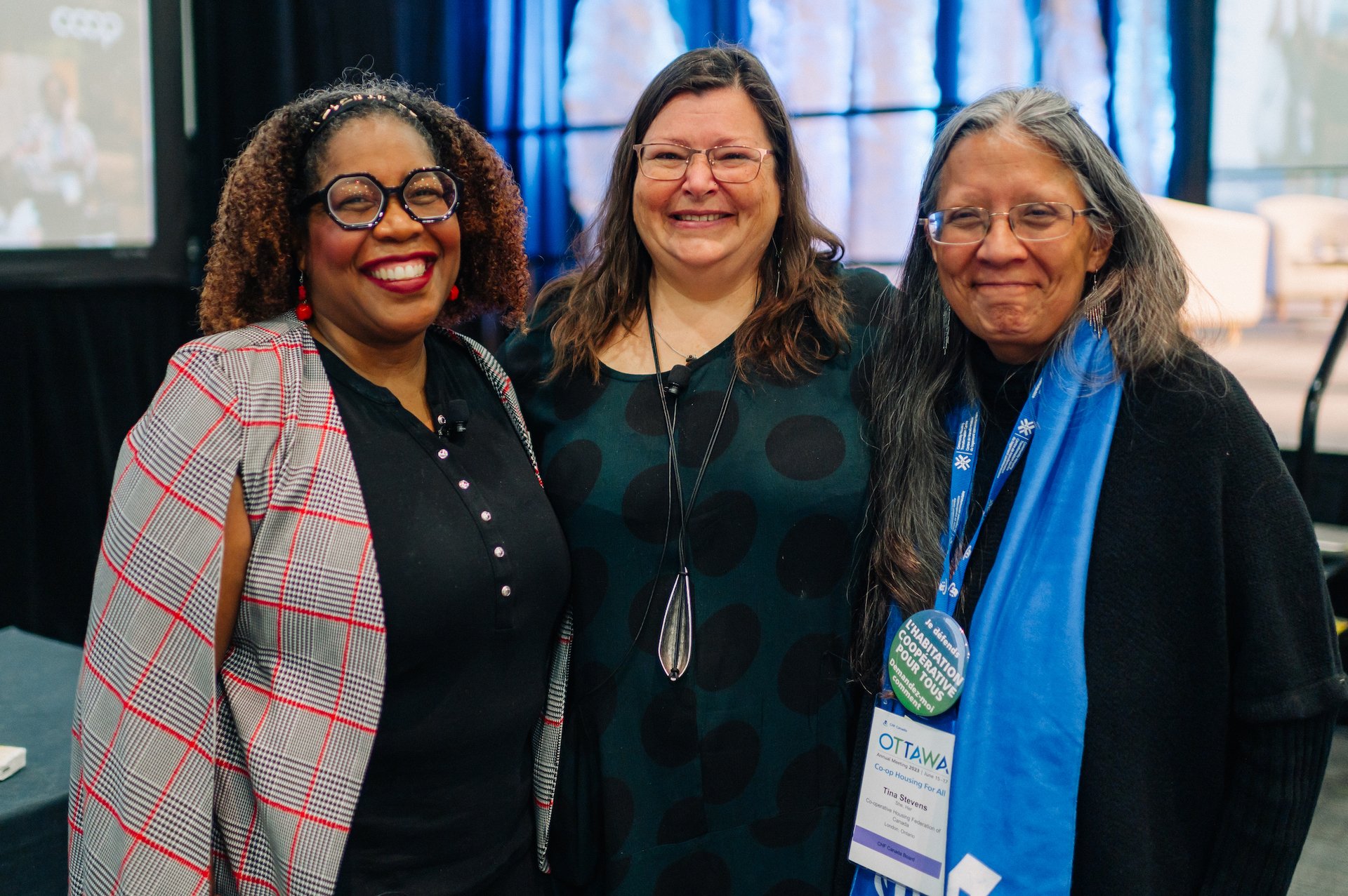 Three women stand side by side, smiling at the camera, wearing conference badges and casual-professional attire at a CHF Canada co-operative housing event.