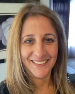A woman with long, straight, light brown hair smiles at the camera indoors, with a framed flower picture and a window in the background—reflecting the warmth of co-operative housing supported by CHF Canada.