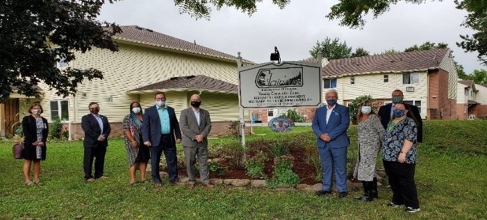 A group of people stands in front of a Co-operative Housing community sign in a residential area, posing for a photo; some are wearing face masks.