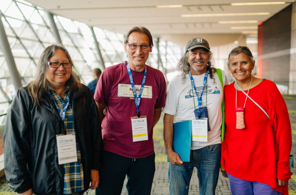 Four adults wearing conference badges stand together indoors, smiling at the camera in a brightly lit, modern venue during a CHF Canada co-operative housing event.